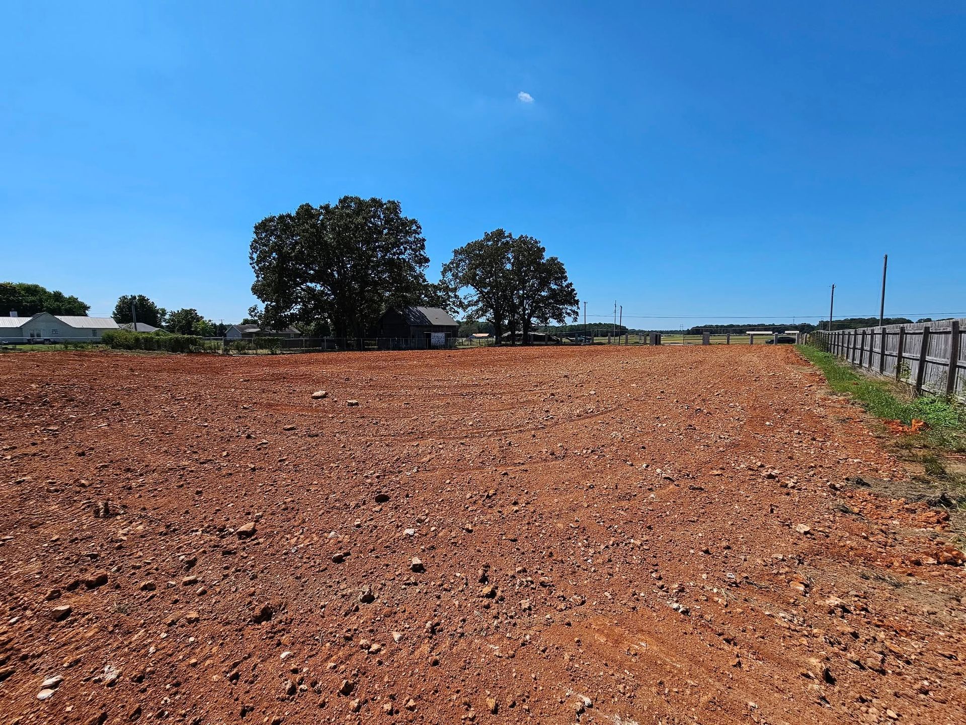 Plowed reddish-brown field under a blue sky, with trees and a fence in the distance.
