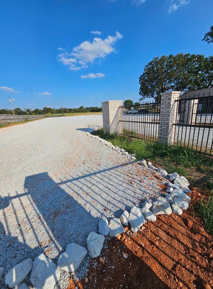 Gravel driveway beside a metal gate and fence under a bright blue sky with clouds.