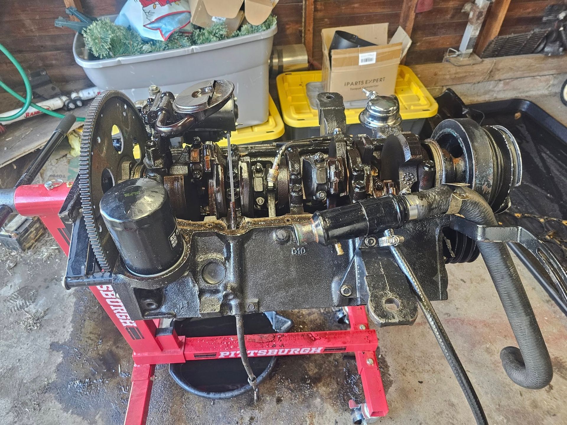 Oil-covered engine on a red stand in a garage, with tools and storage bins in the background.