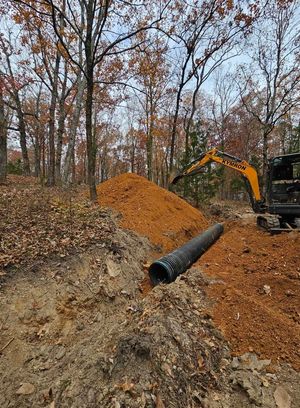Excavator digging in a wooded area beside a dirt mound and black drainage pipe.