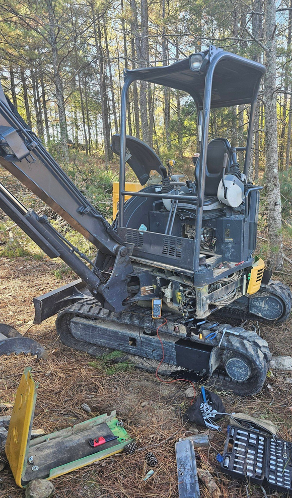 A small excavator with its bucket arm extended, parked in a wooded area with tools on the ground.