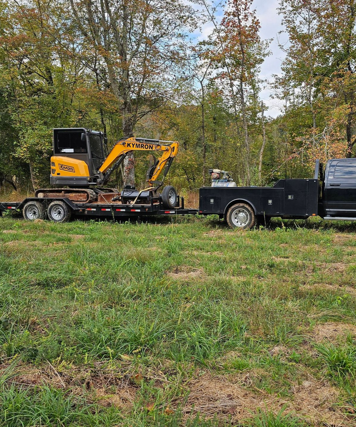 Yellow excavator on a trailer hitched to a black truck in a grassy field with trees behind.