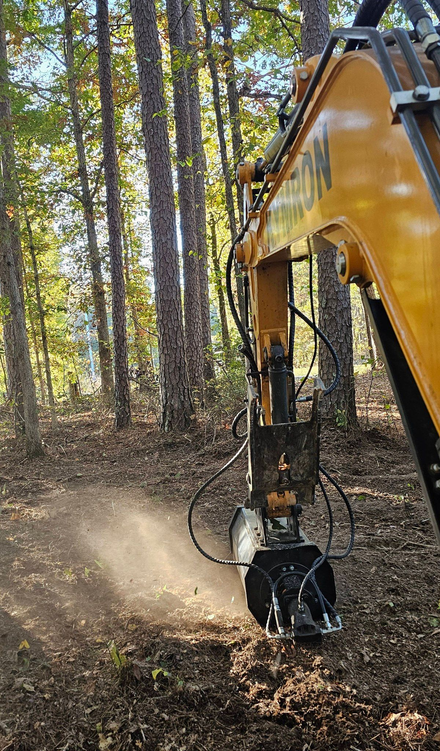 Excavator mulching brush in a wooded area, kicking up dust among tall trees.