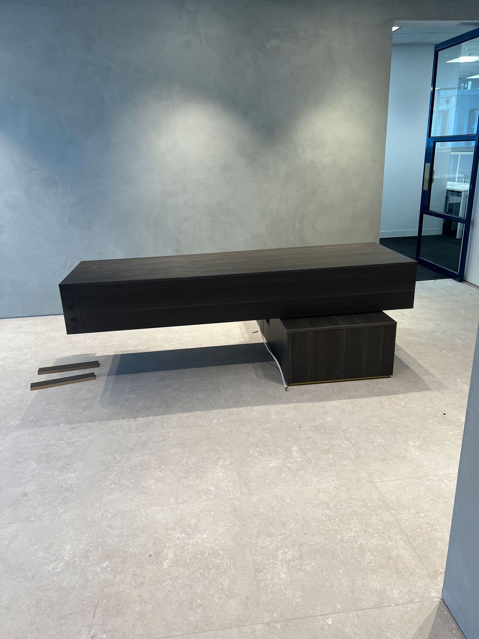 Dark, rectangular table floating above a smaller cube-shaped base in a room with light gray walls and floor.