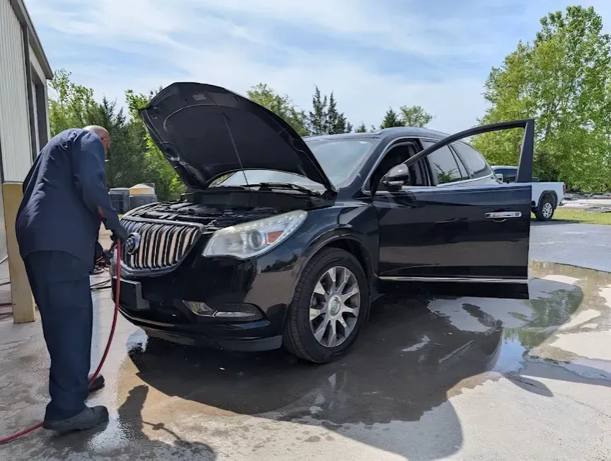 Man washing a black SUV with hood and driver door open on a driveway, water pooling nearby.