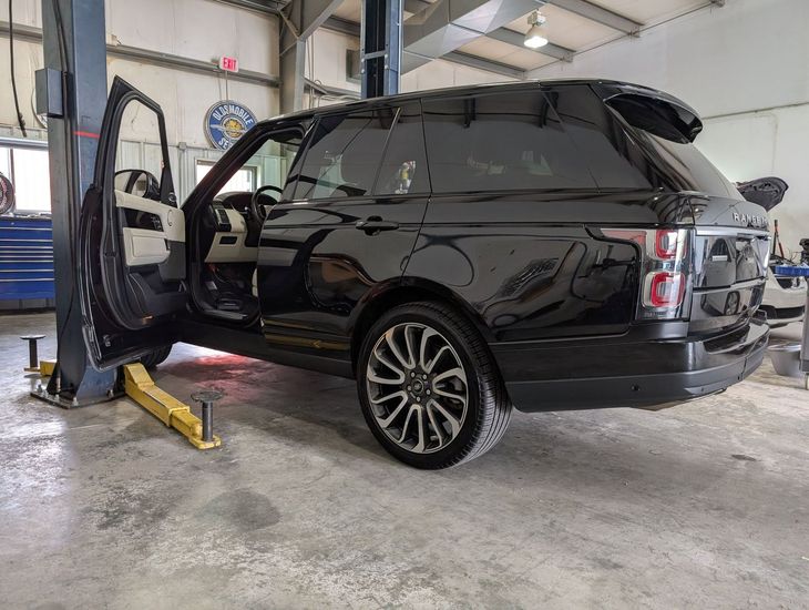 Black SUV with open driver door in a garage, rear panel lifted and rear wheel visible on a lift.