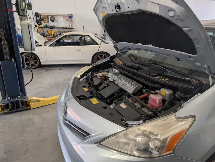 Silver car with hood open in an auto repair shop, another white car on a lift in the background