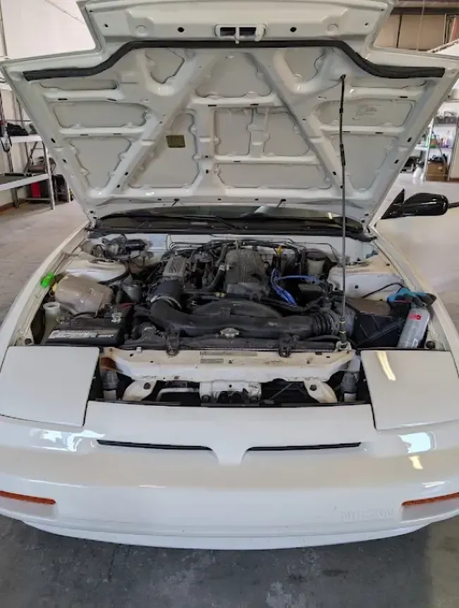 White car with hood open, showing the engine bay in a garage.