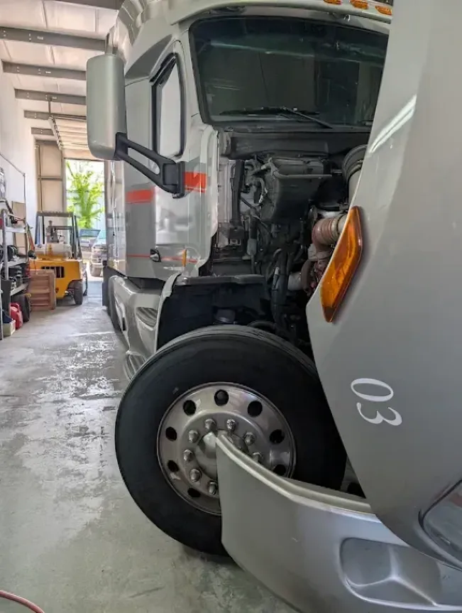 Two trucks parked in a garage, one silver truck with its front panel open exposing the engine.