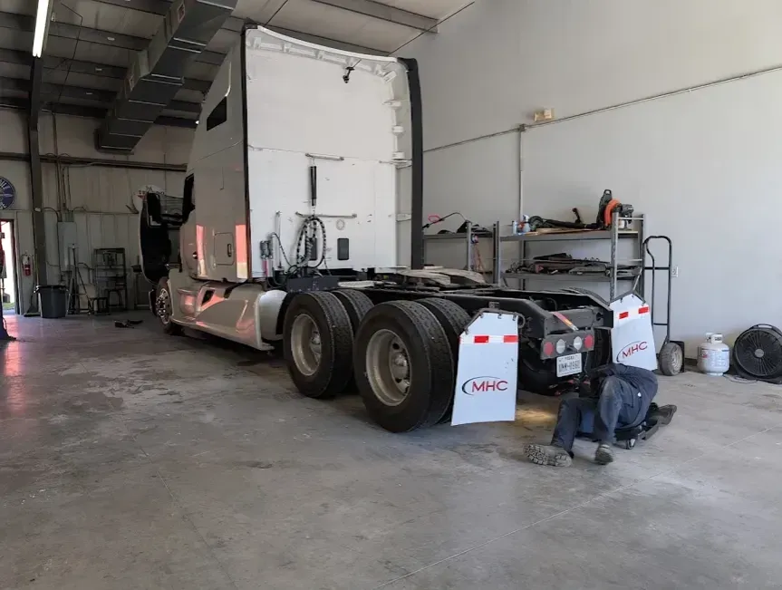 White semi-truck chassis in a garage with a person crouching beside the rear wheel.