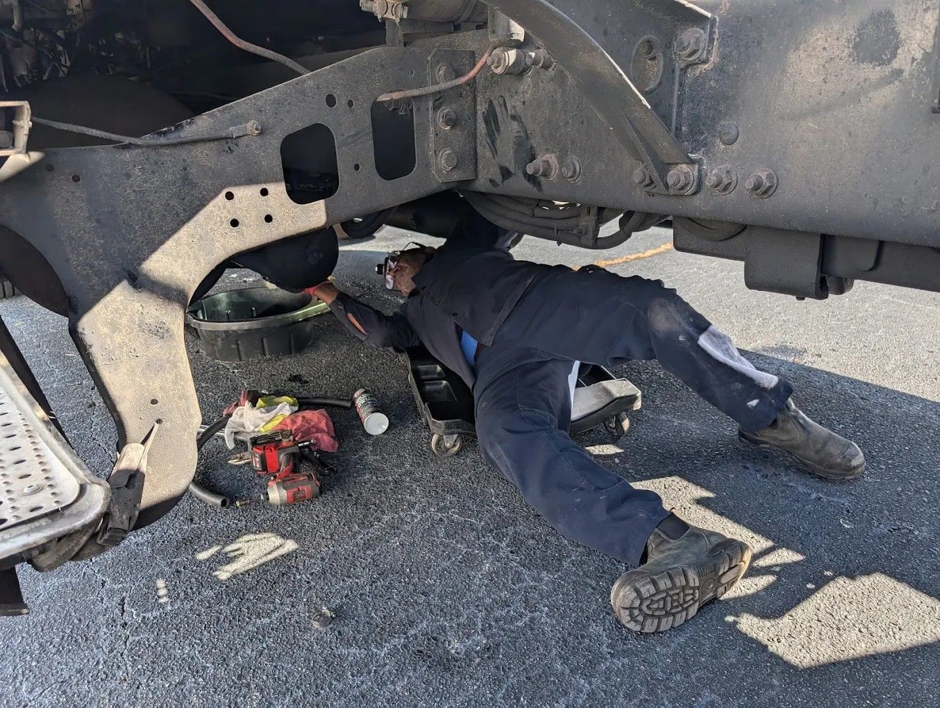 Person lying under a damaged vehicle on a gravel road beside debris and a red object