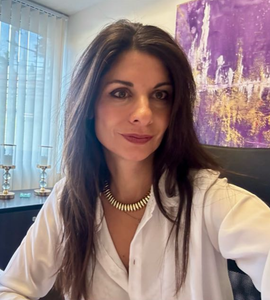 Mujer con cabello castaño y collar dorado con camisa blanca, sentada en un escritorio