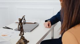 Escritorio con estatua de Lady Justice, computadora portátil, papeles y dos personas, una de ellas con un blazer azul.