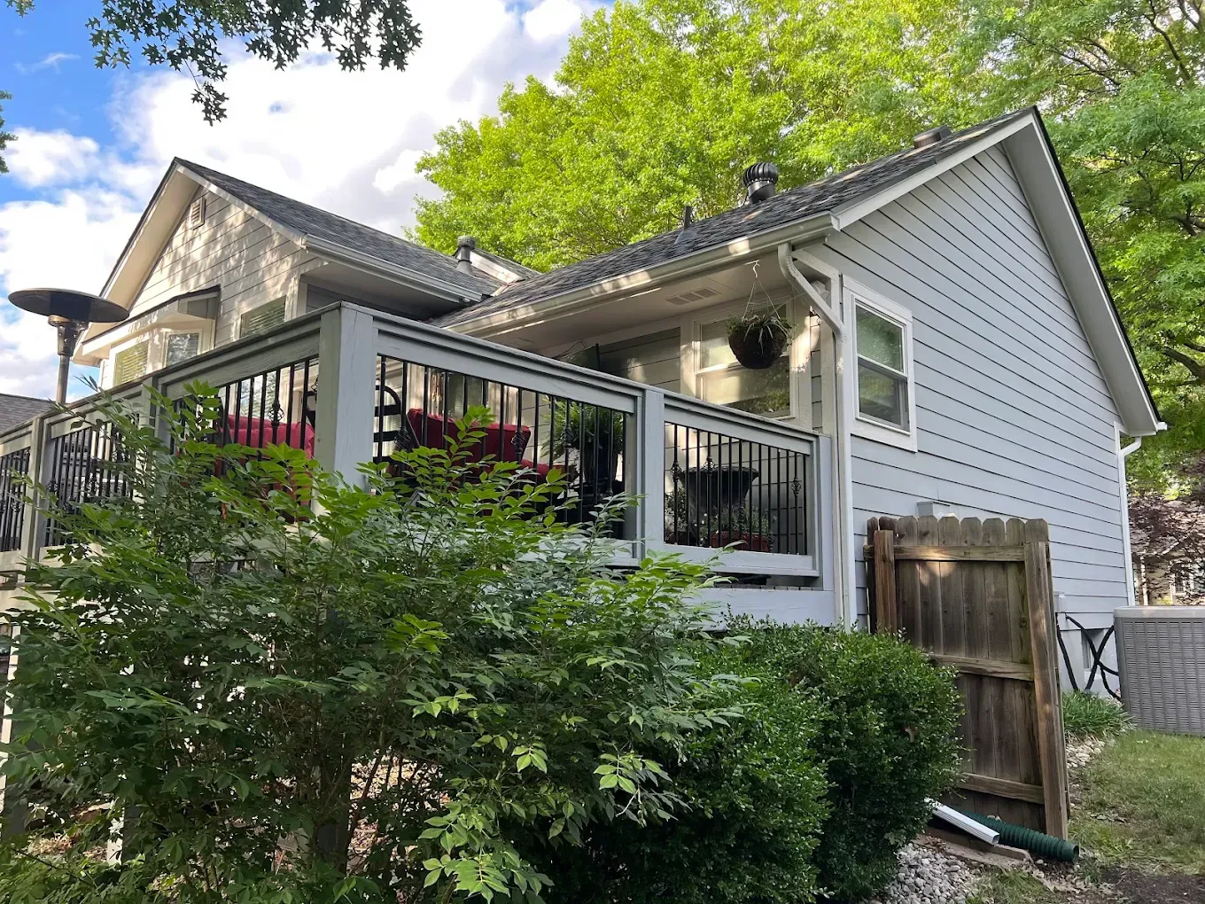 A side view of a gray-sided house featuring a raised wooden deck with black railings, surrounded by green landscaping.
