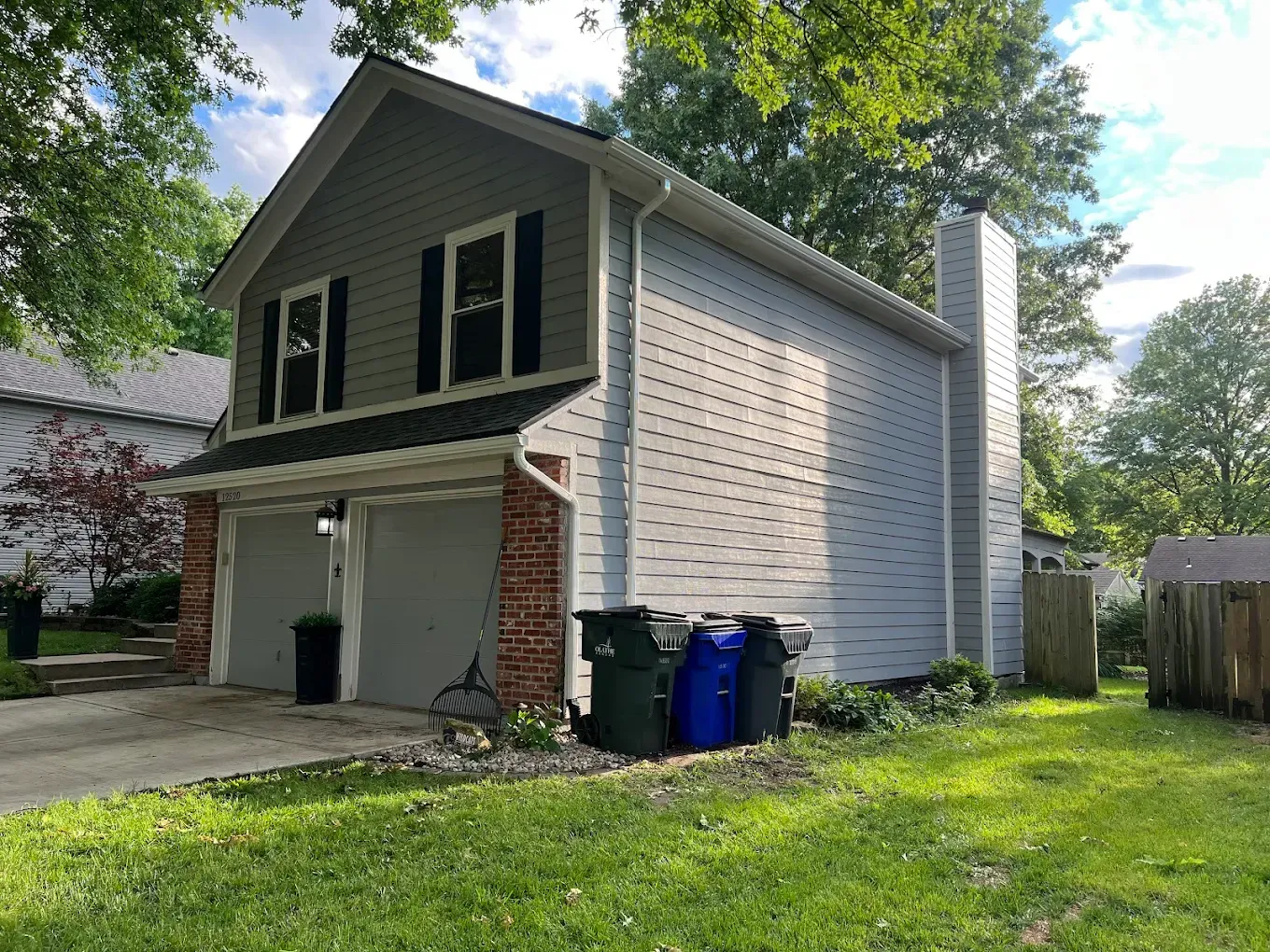 A two-story garage with grey siding, brick trim around the doors, black shutters, and three trash bins outside.
