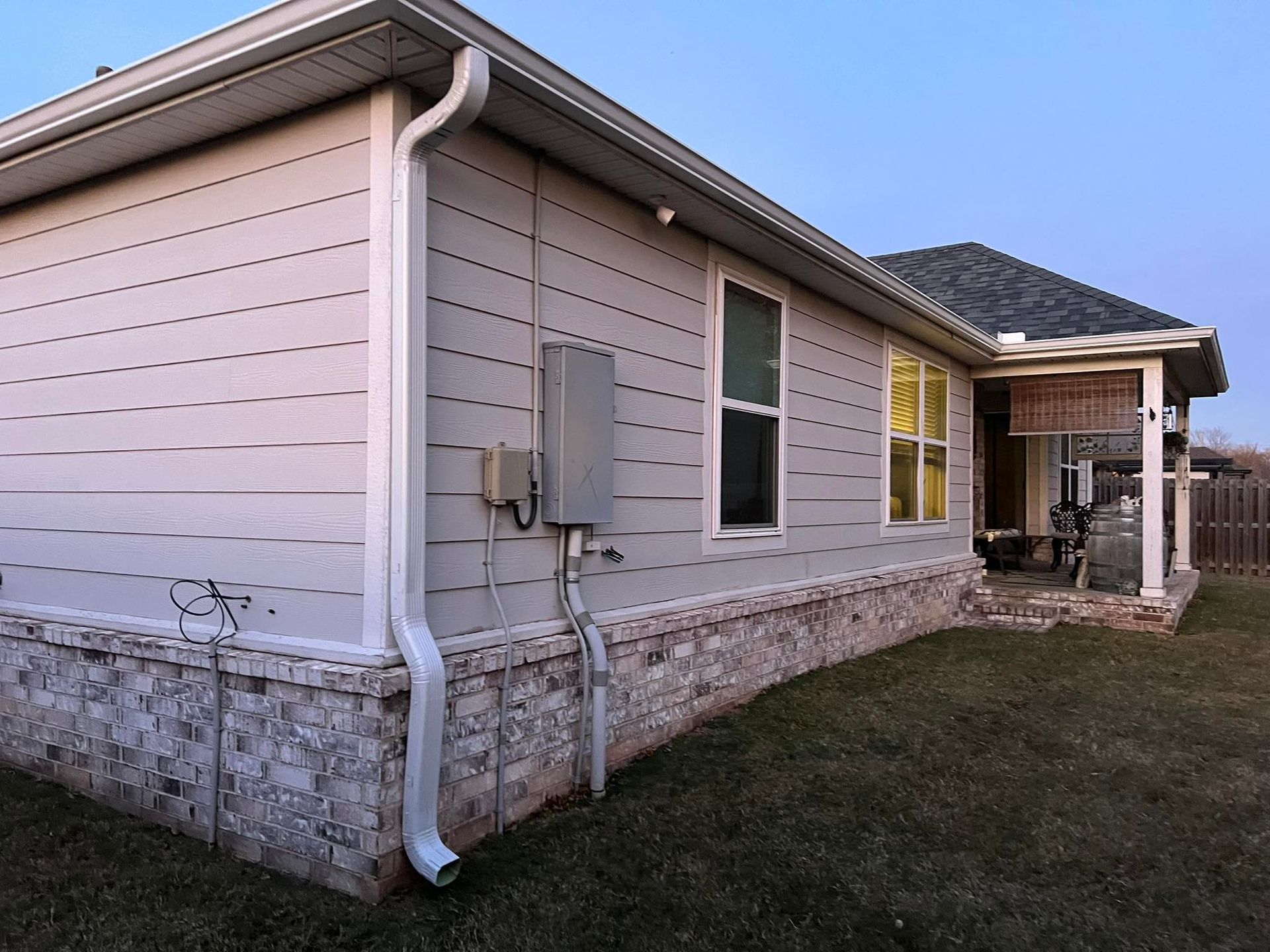 The side of a house with light gray horizontal siding, a stone foundation, a white gutter, and an electrical box.