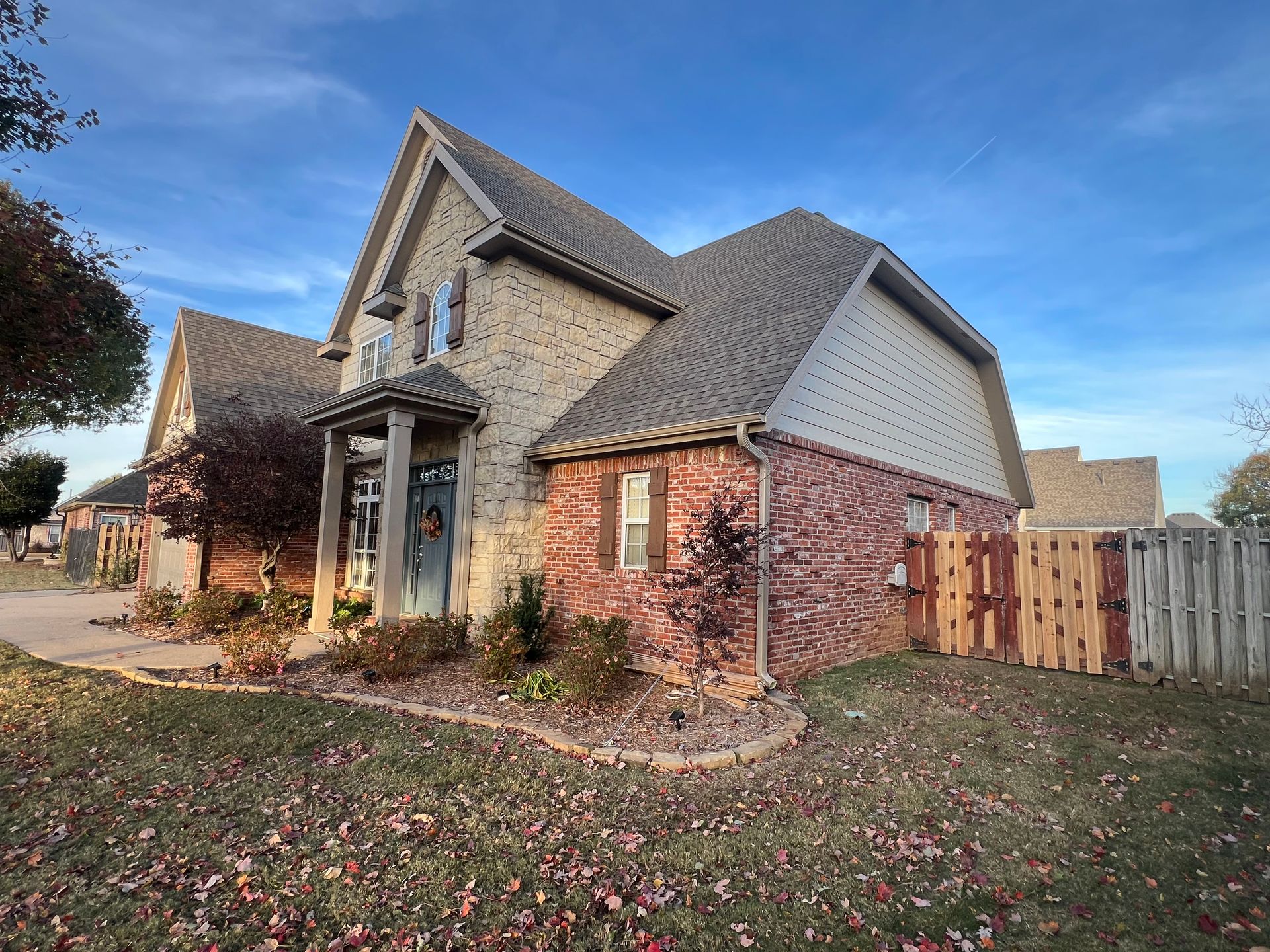 A two-story house with a brick exterior, stone entryway, and steep shingled roof, surrounded by a lawn with fallen leaves.