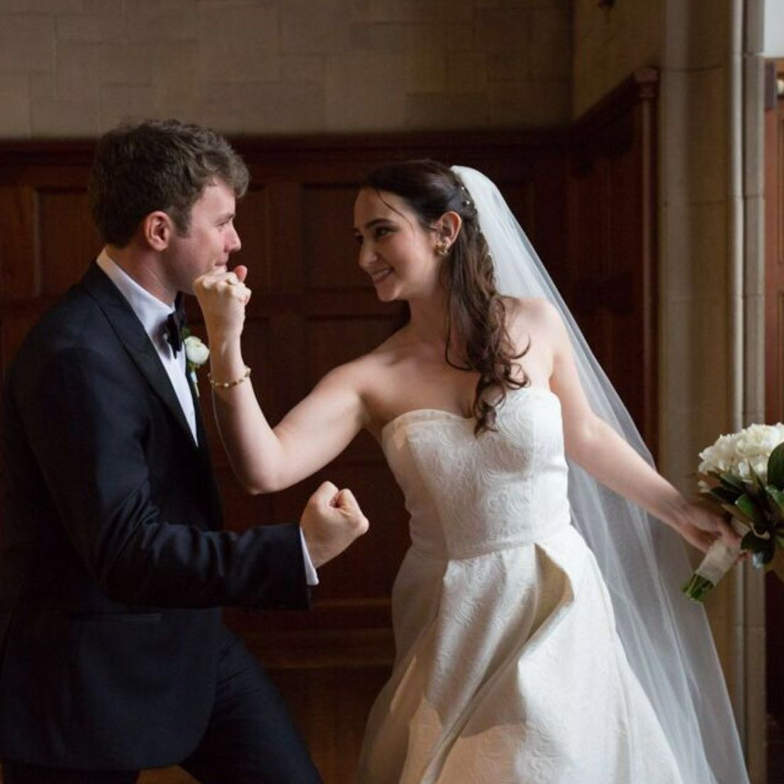 Bride and groom smiling in a wood-paneled room, holding hands during their wedding dance.