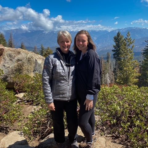 Two people standing on a mountain overlook with trees, rocks, and distant peaks under a blue sky.