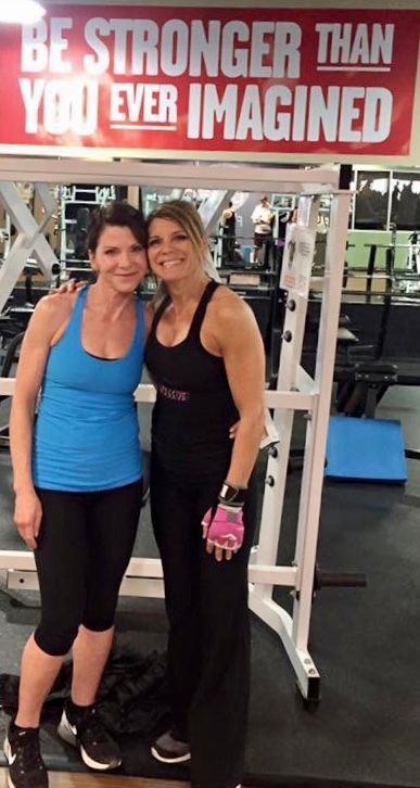 Two women posing in a gym under a “Be Stronger Than You Ever Imagined” sign.