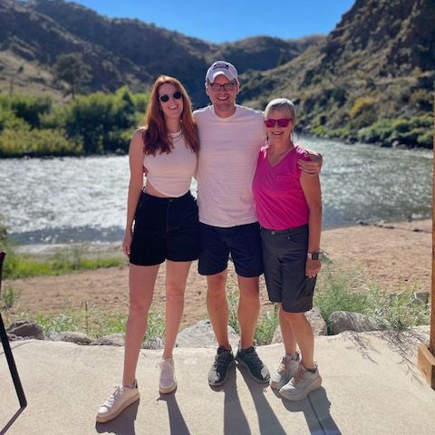 Three people posing by a river with mountains in the background on a sunny day