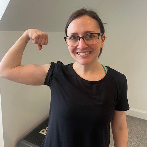 Person smiling and flexing an arm in a black shirt, standing indoors against a light wall.