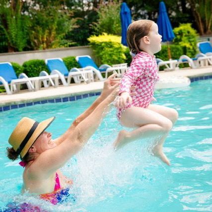 Adult in sunhat lifts child in pink swimsuit into a pool with lounge chairs and umbrellas in the background