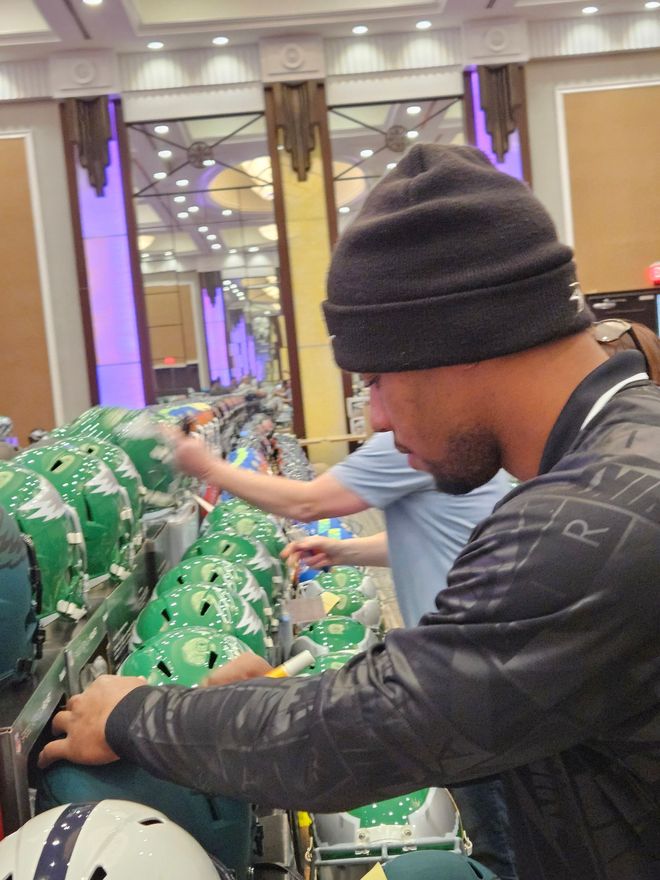 Man in black beanie working on football helmets. Green and white helmets in a well-lit room with gold accents.