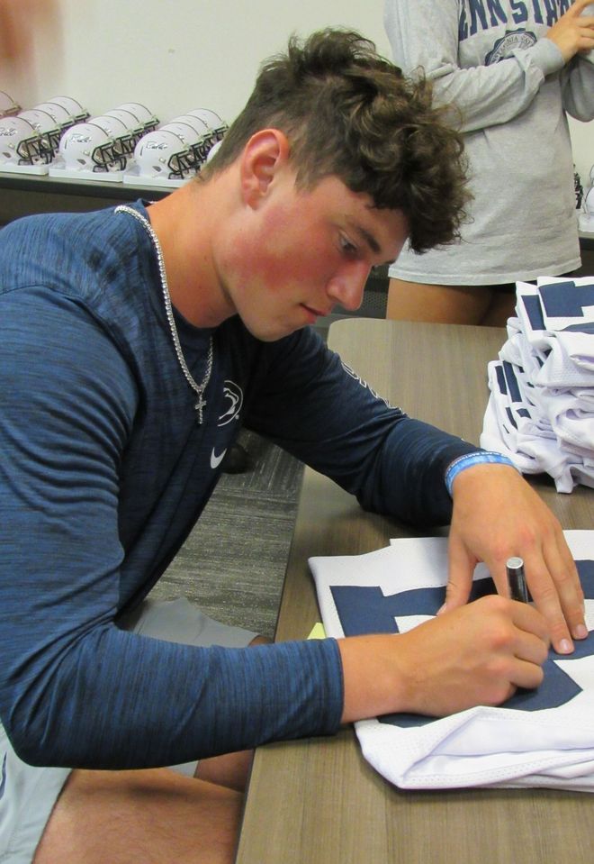 Man with curly hair signing a jersey. He's wearing a blue shirt and necklace. White jerseys stacked on the table.