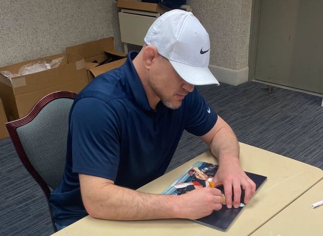 Man in white cap signing something on a table. He wears a navy polo shirt and is indoors.