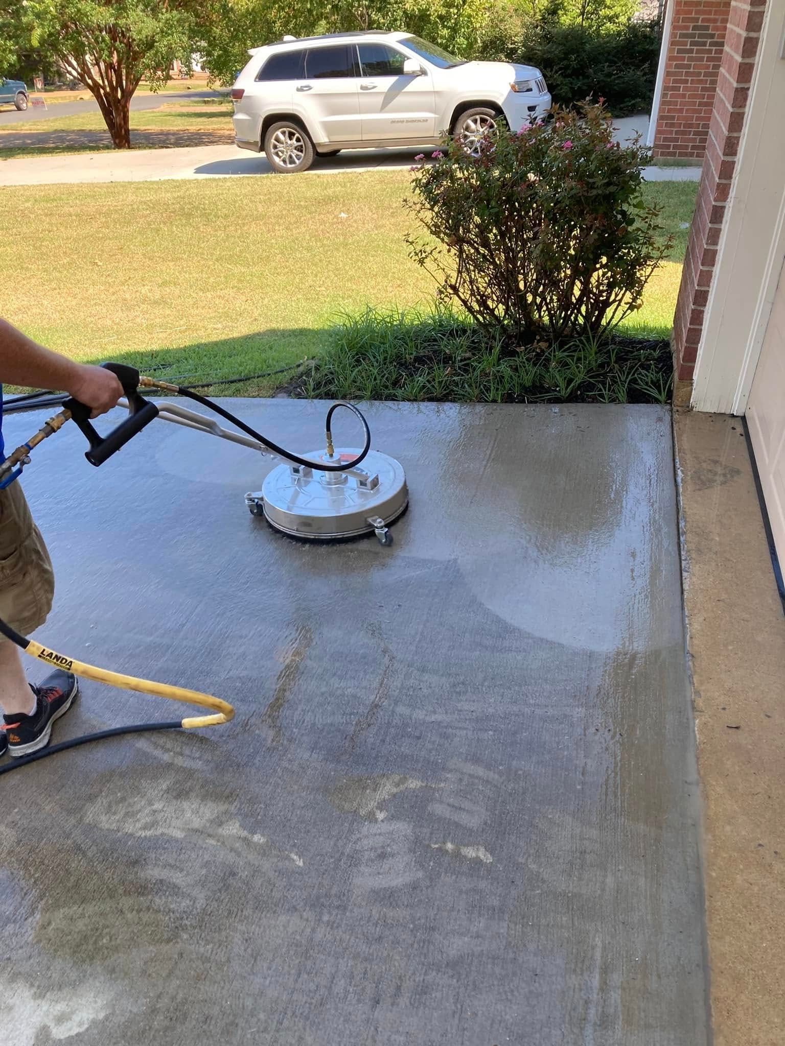 A person uses a circular surface cleaner attachment with a pressure washer on a concrete driveway.