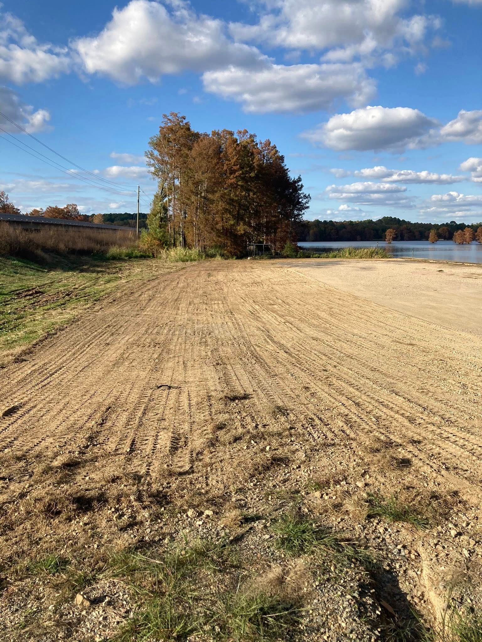 A freshly plowed dirt field under a bright blue, cloudy sky, with a cluster of trees near a body of water in the distance.