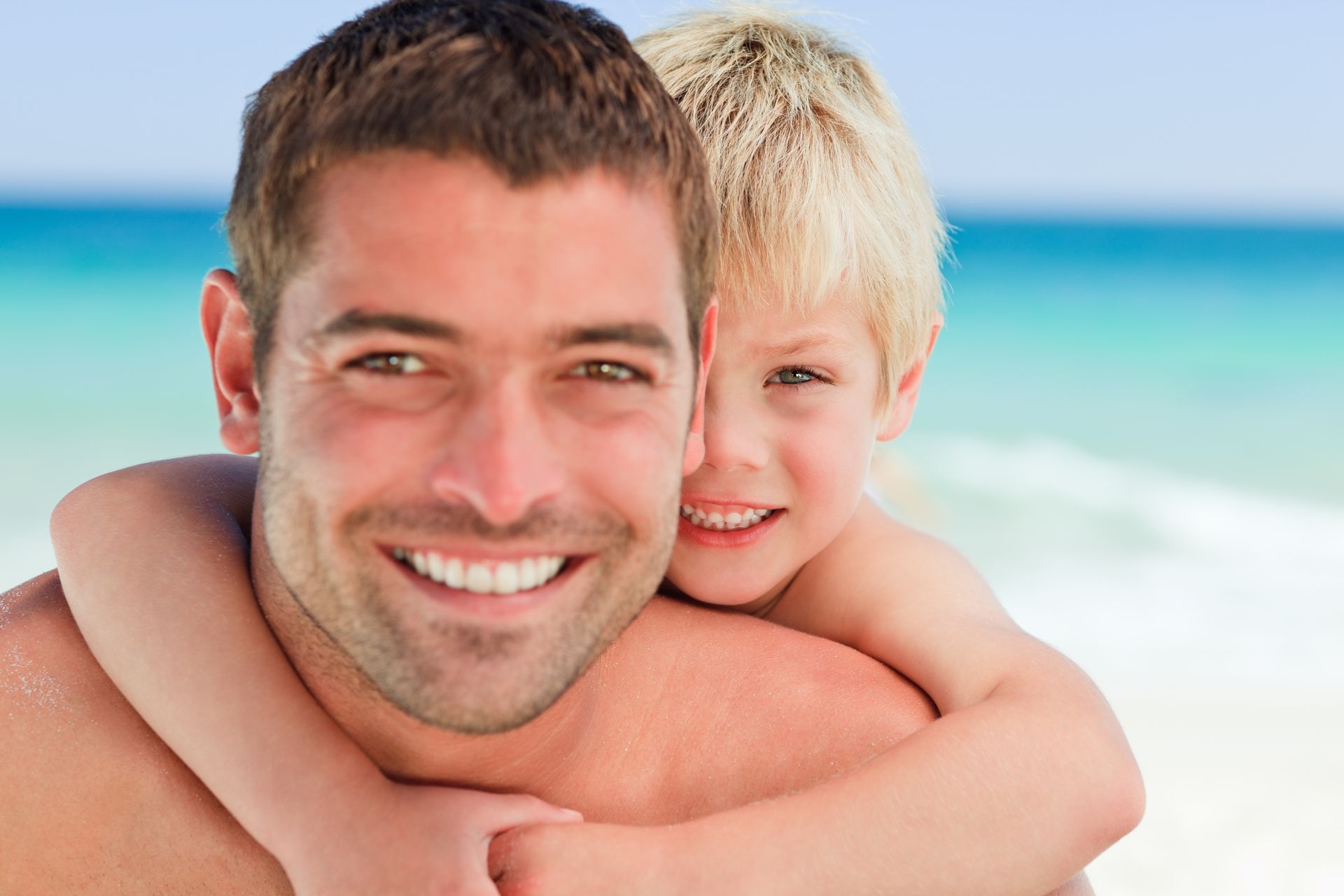 A smiling person giving a piggyback ride to a child on a sunny beach.
