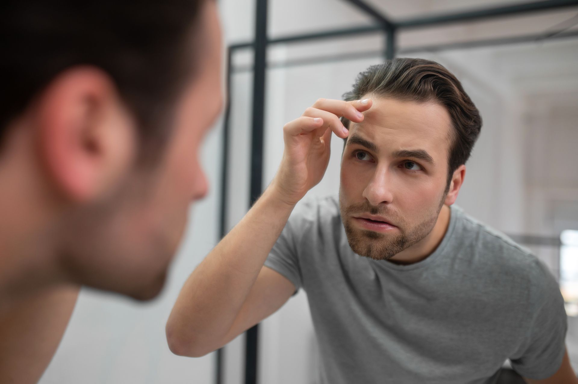 A person in a gray t-shirt looks into a mirror, touching their forehead while examining their face.
