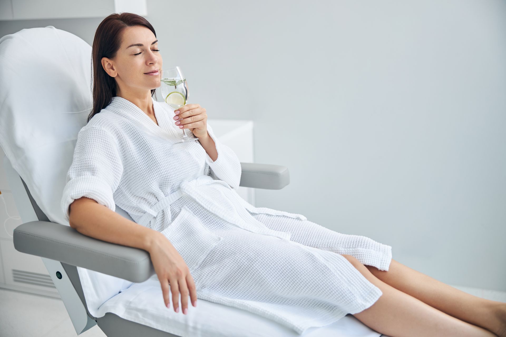A person in a white waffle-weave robe rests in a spa chair, eyes closed, holding a glass with a lime wedge.