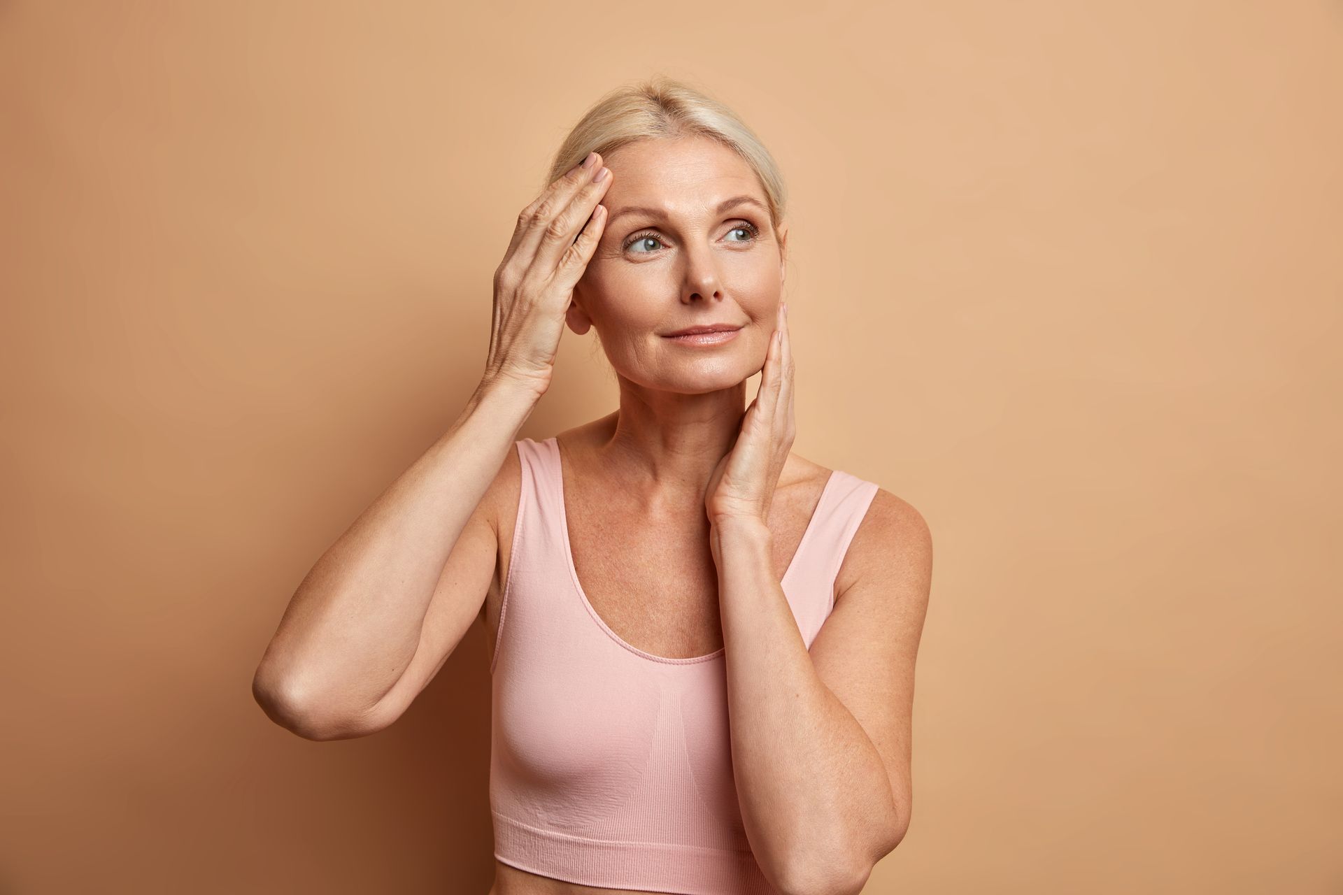 A person in a light pink tank top touches their face while looking away against a plain tan background.