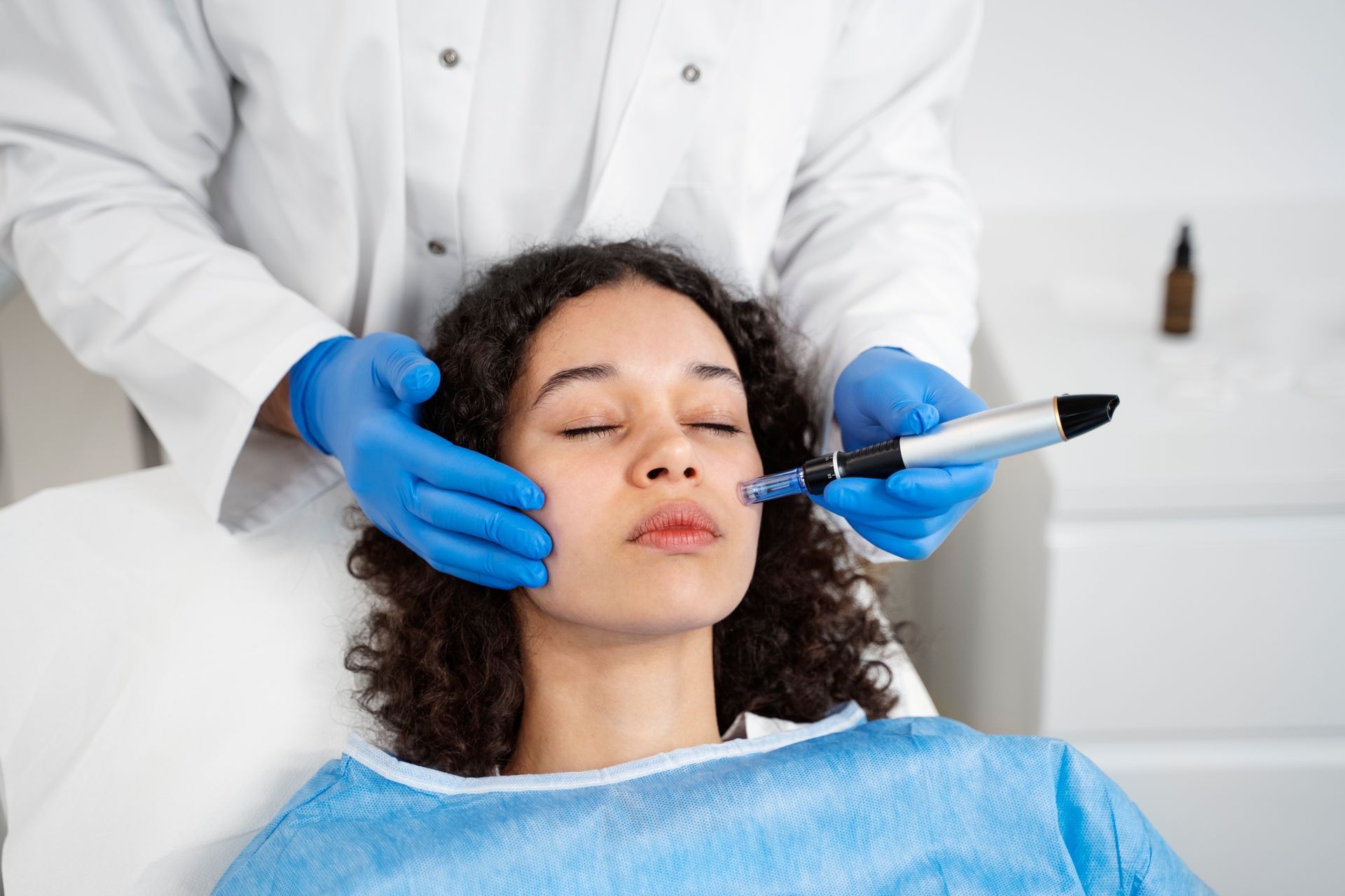 A practitioner in white medical attire and blue gloves performs a microneedling procedure on a person's face.