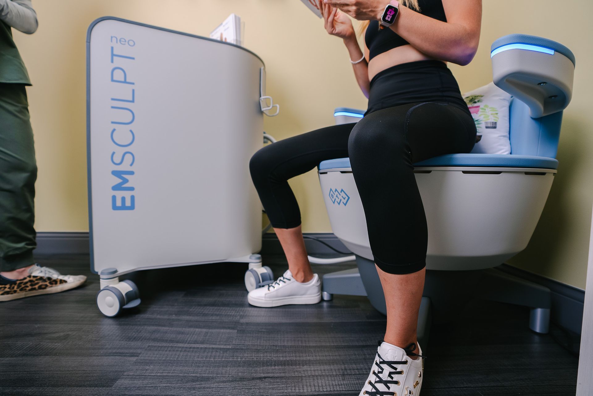 Person seated on an Emsculpt machine in a clinic, wearing black workout clothes and sneakers.