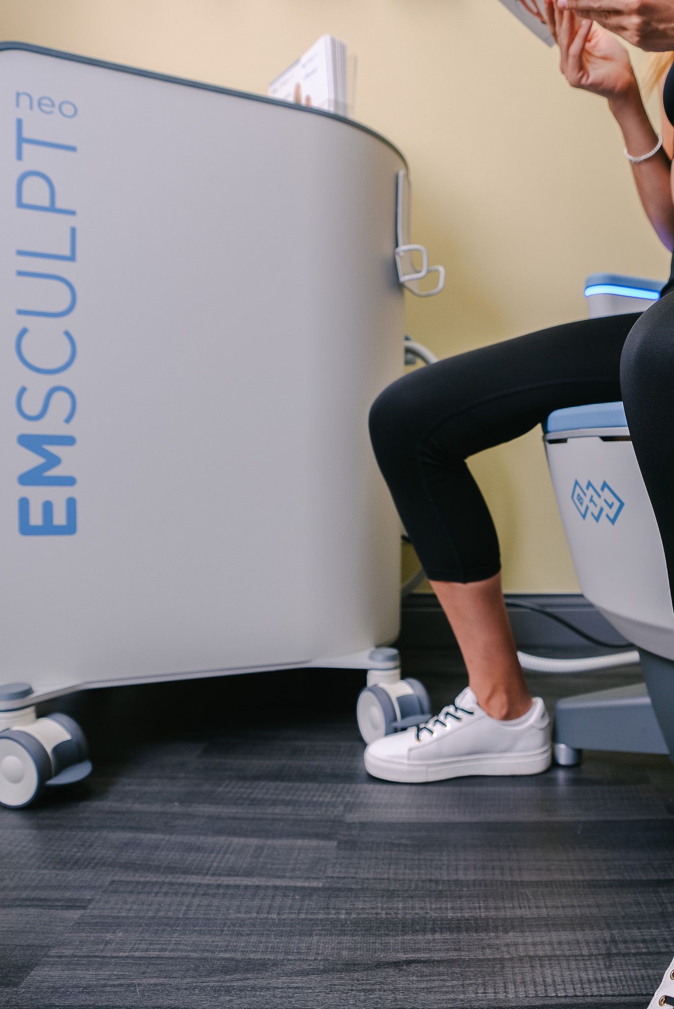 Person seated on an Emsculpt Neo machine in a clinic, wearing black leggings and white sneakers.