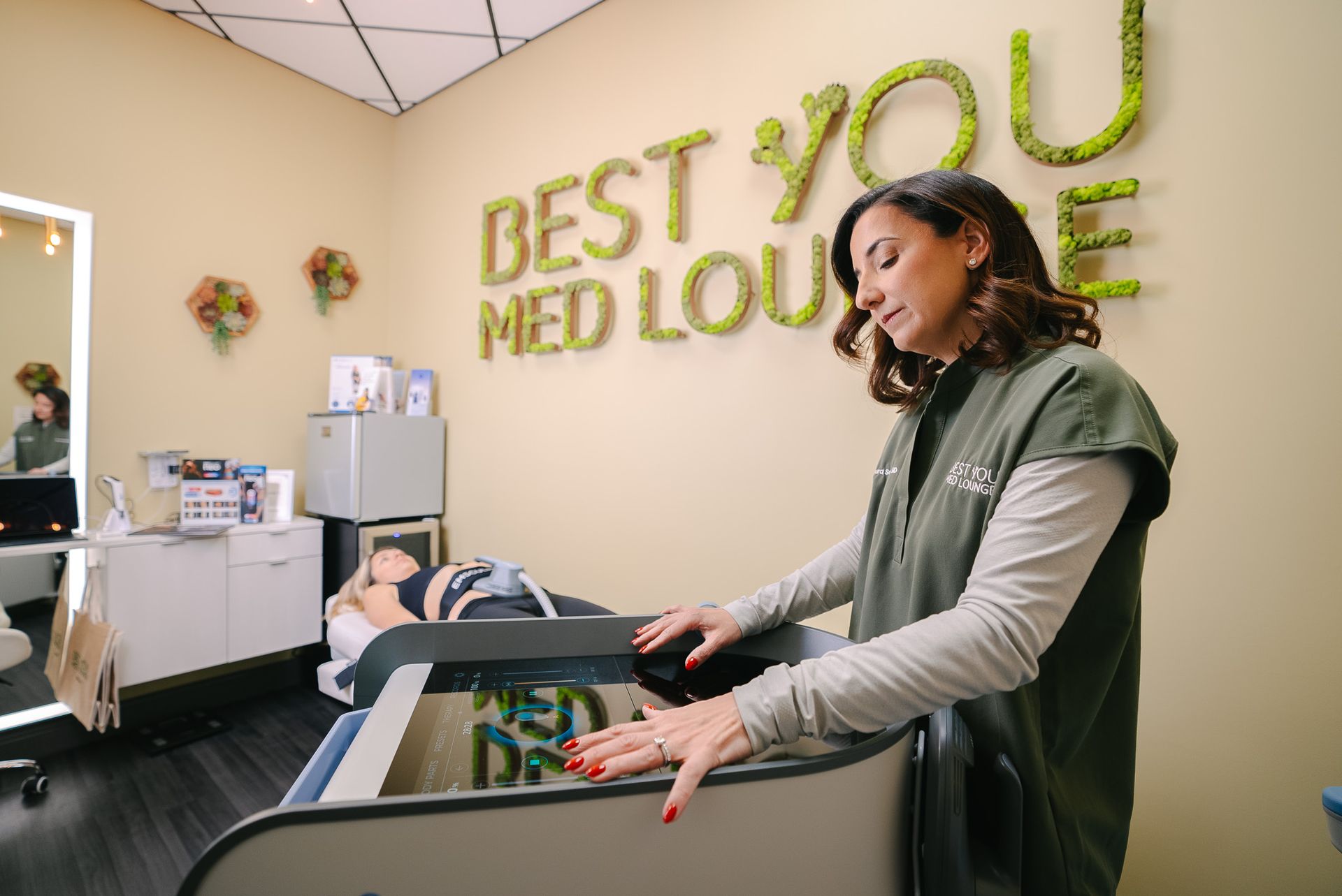 Woman at a medical spa reception desk with green “BEST YOU MED LOUNGE” wall sign