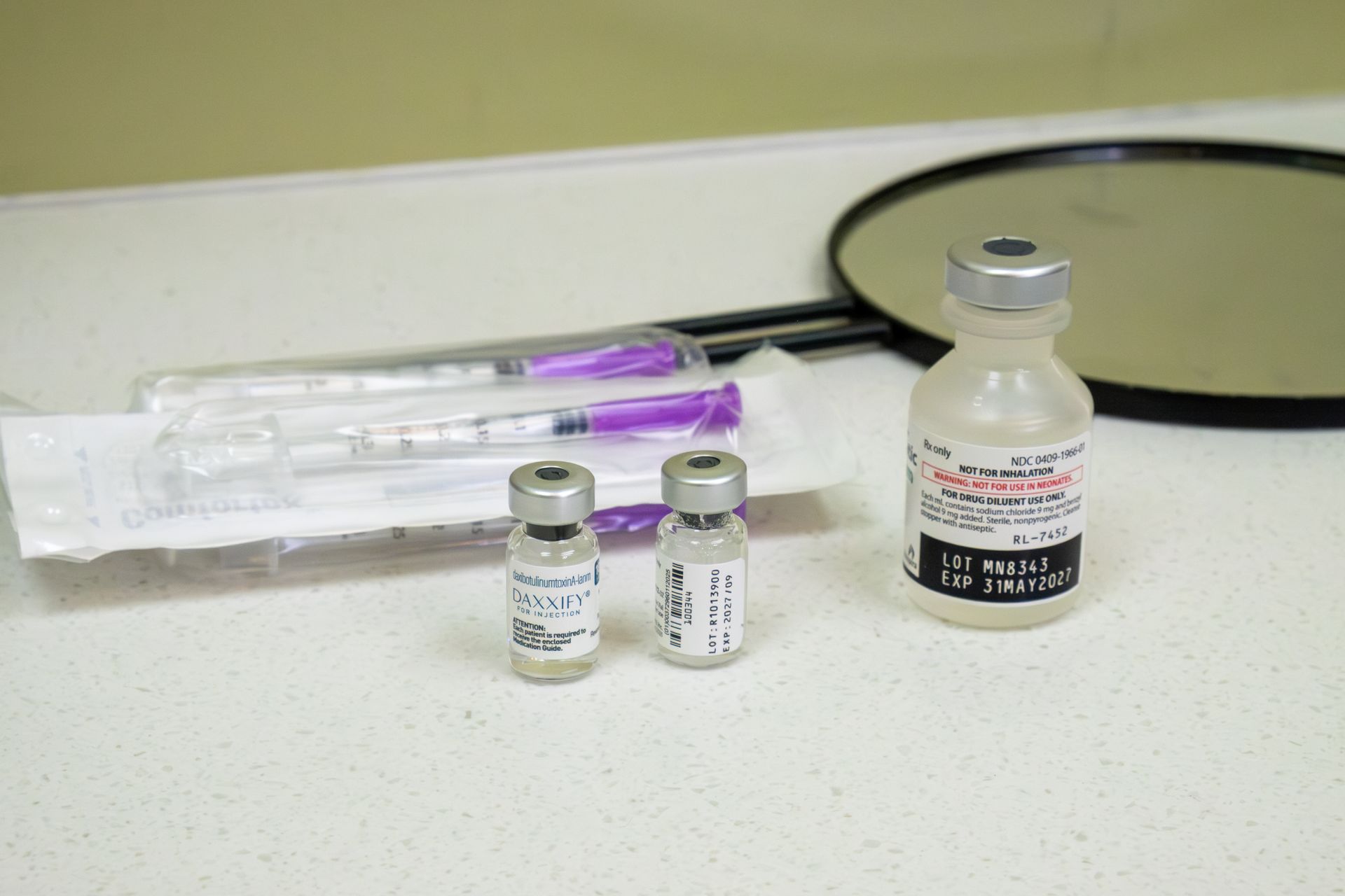 Medical vials and a syringe on a countertop beside a round mirror
