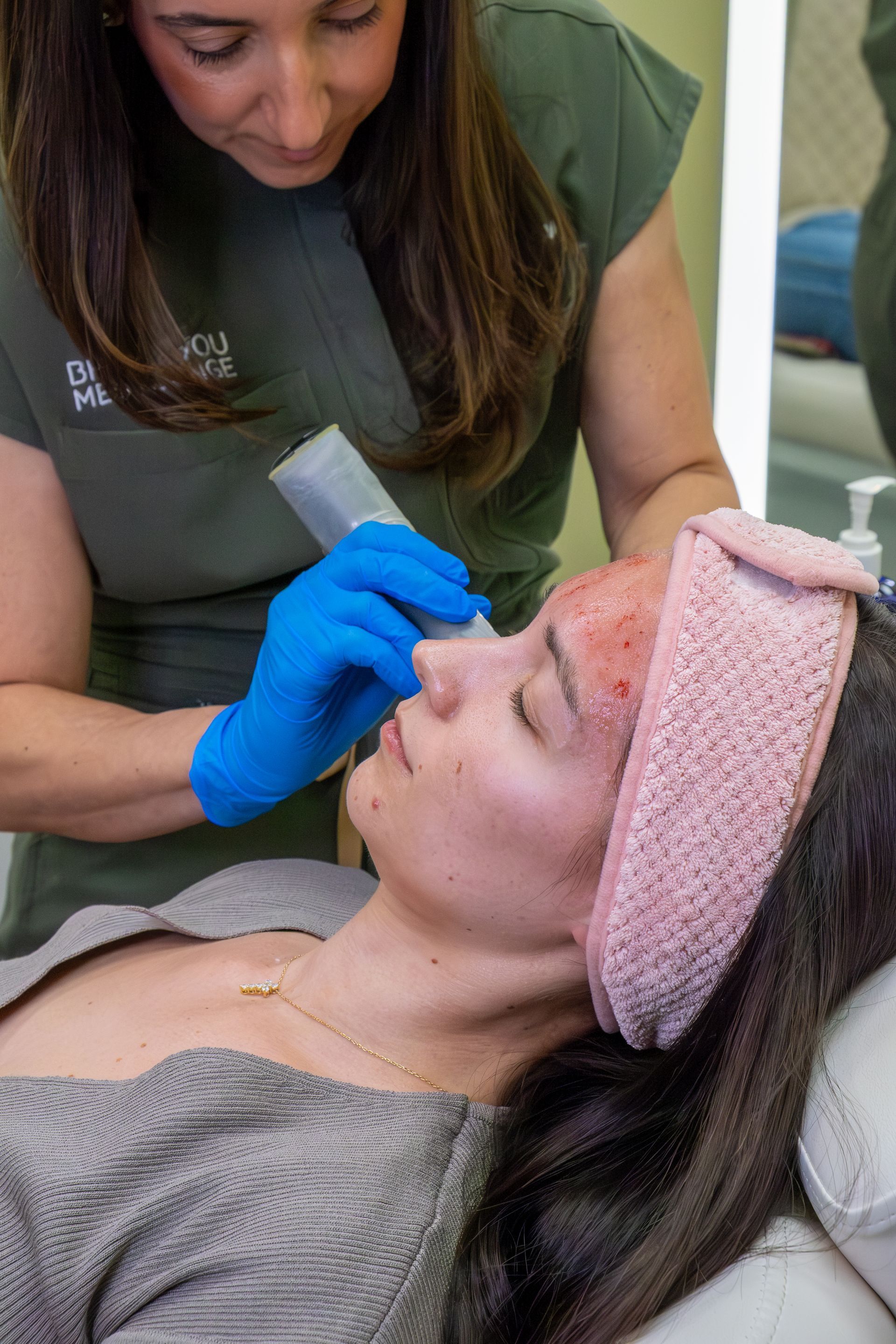 Esthetician applying treatment to a client’s face while she reclines with a pink headband