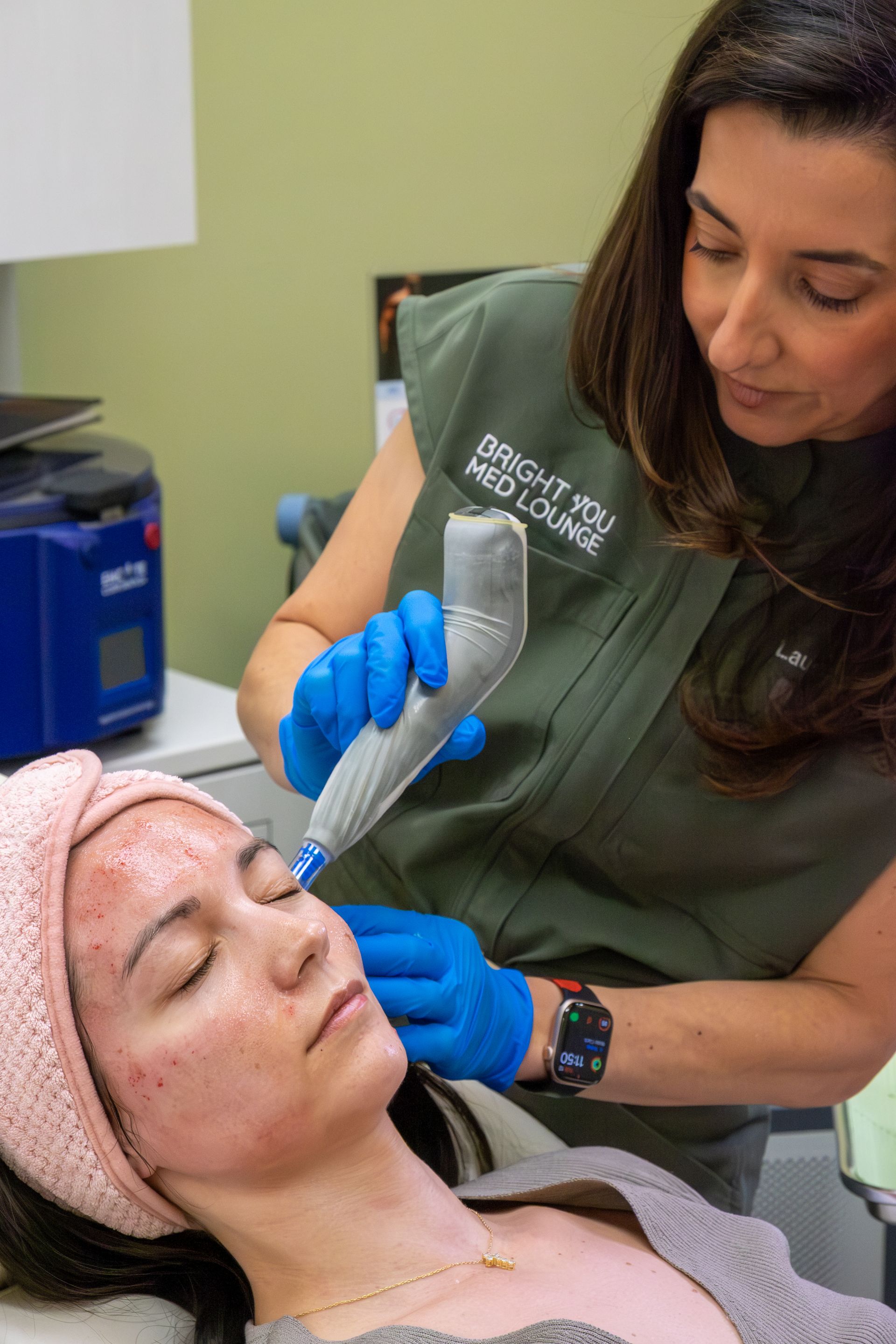 A clinician in blue gloves treats a client’s forehead with a handheld device in a spa setting.