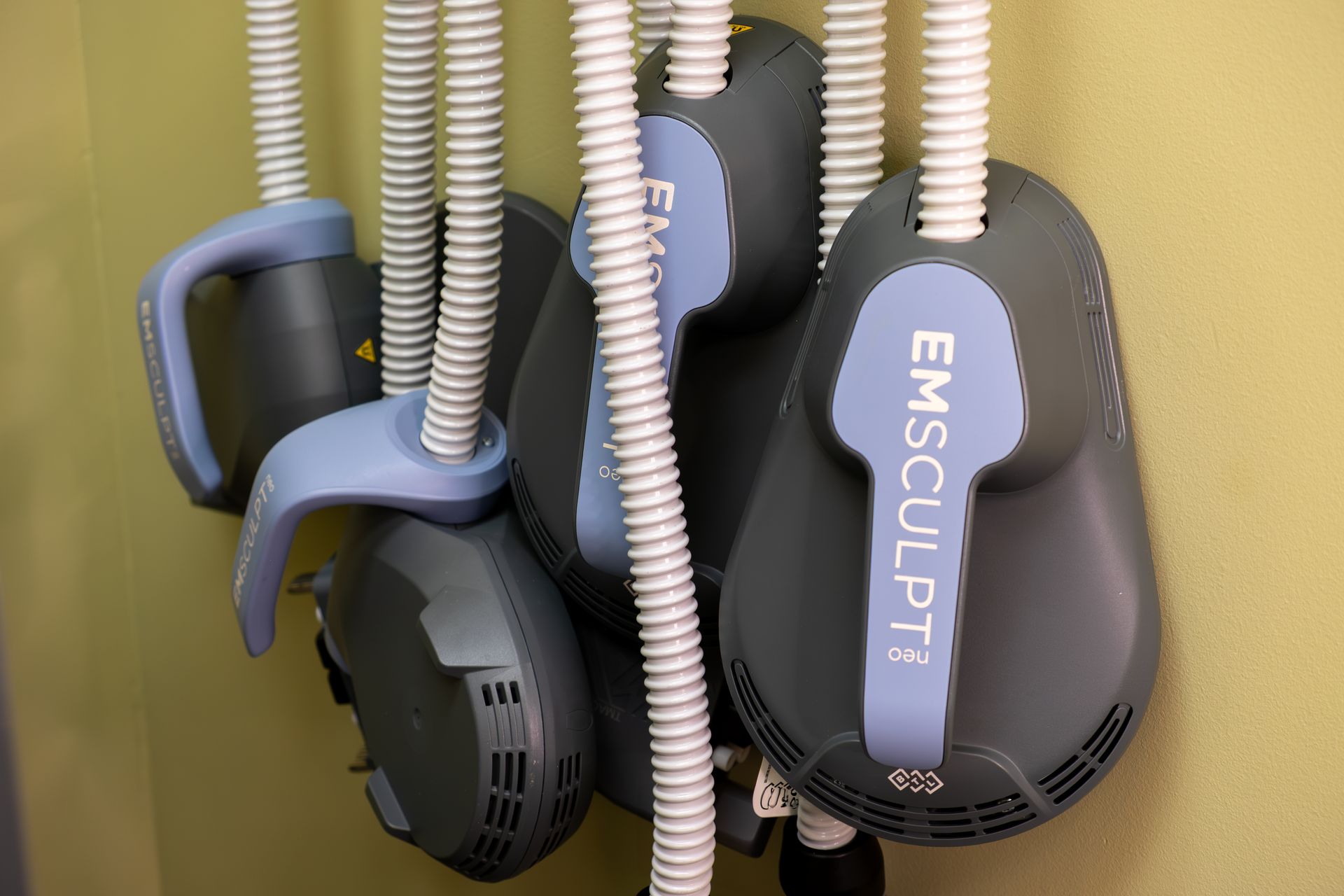 Gym devices labeled EMSculpt with blue handles and black straps hanging on a wall rack