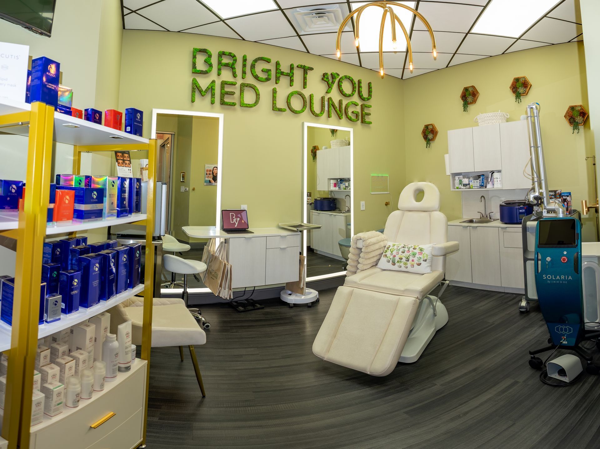 Interior of Bright You Med Lounge: beige medical chair, skincare products on shelves, green and gold decor.
