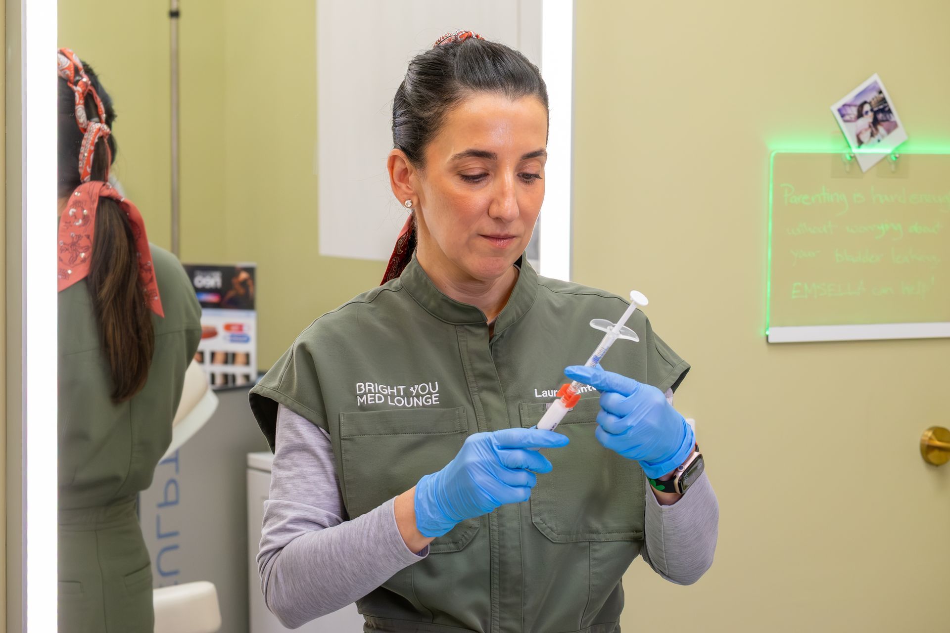 Person in a green vest and blue gloves prepares a syringe in a clinical room.