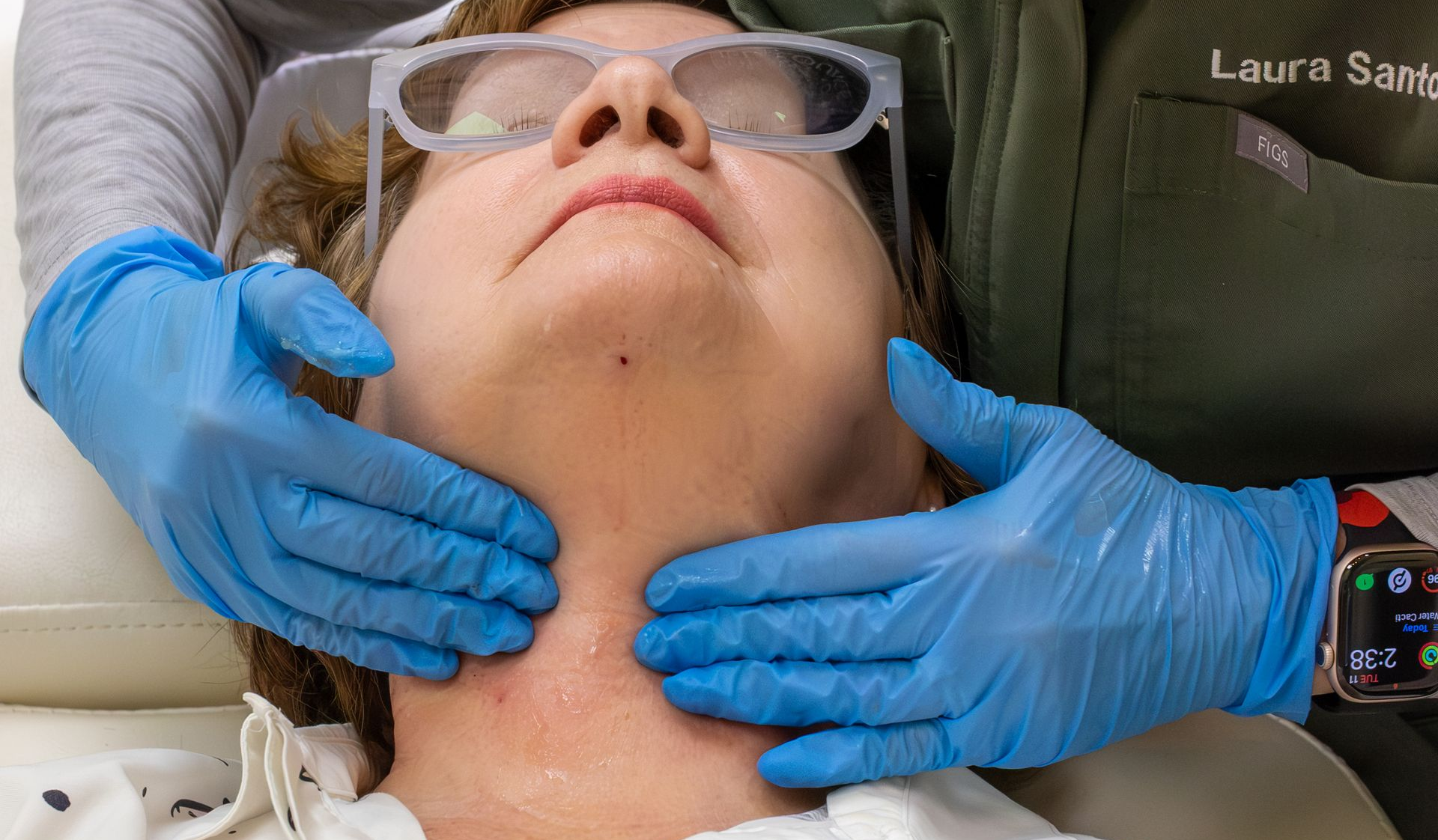Gloved hands massage a patient’s neck while she reclines with eyes closed and protective goggles on.