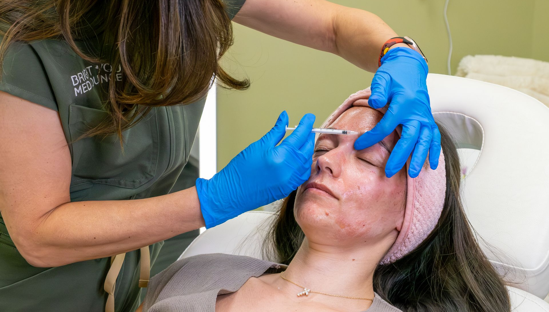Esthetician applying facial treatment to a client lying on a spa bed, wearing blue gloves and a pink headband