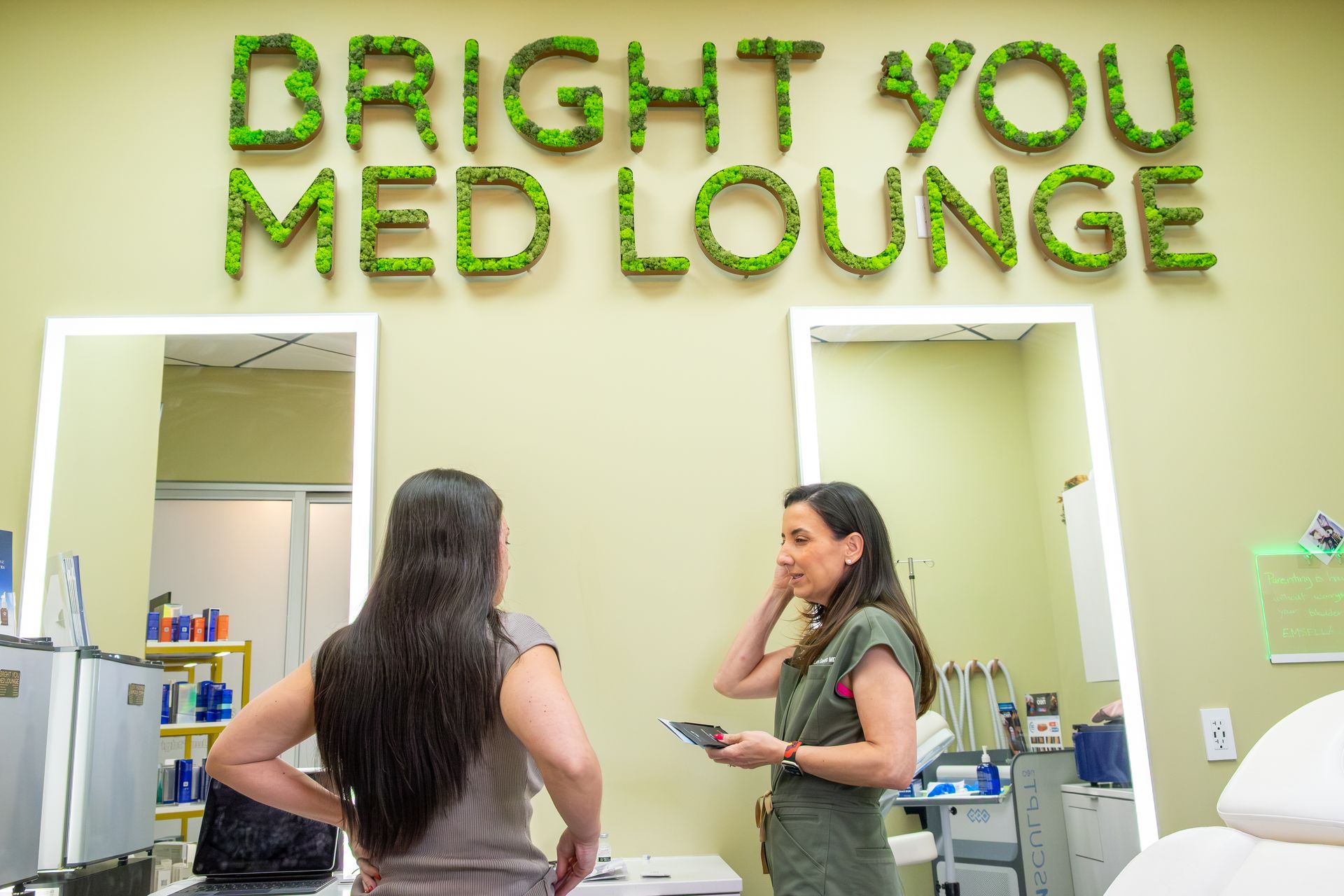 Two people talk at the front desk of Bright You Med Lounge with green wall signage behind them.