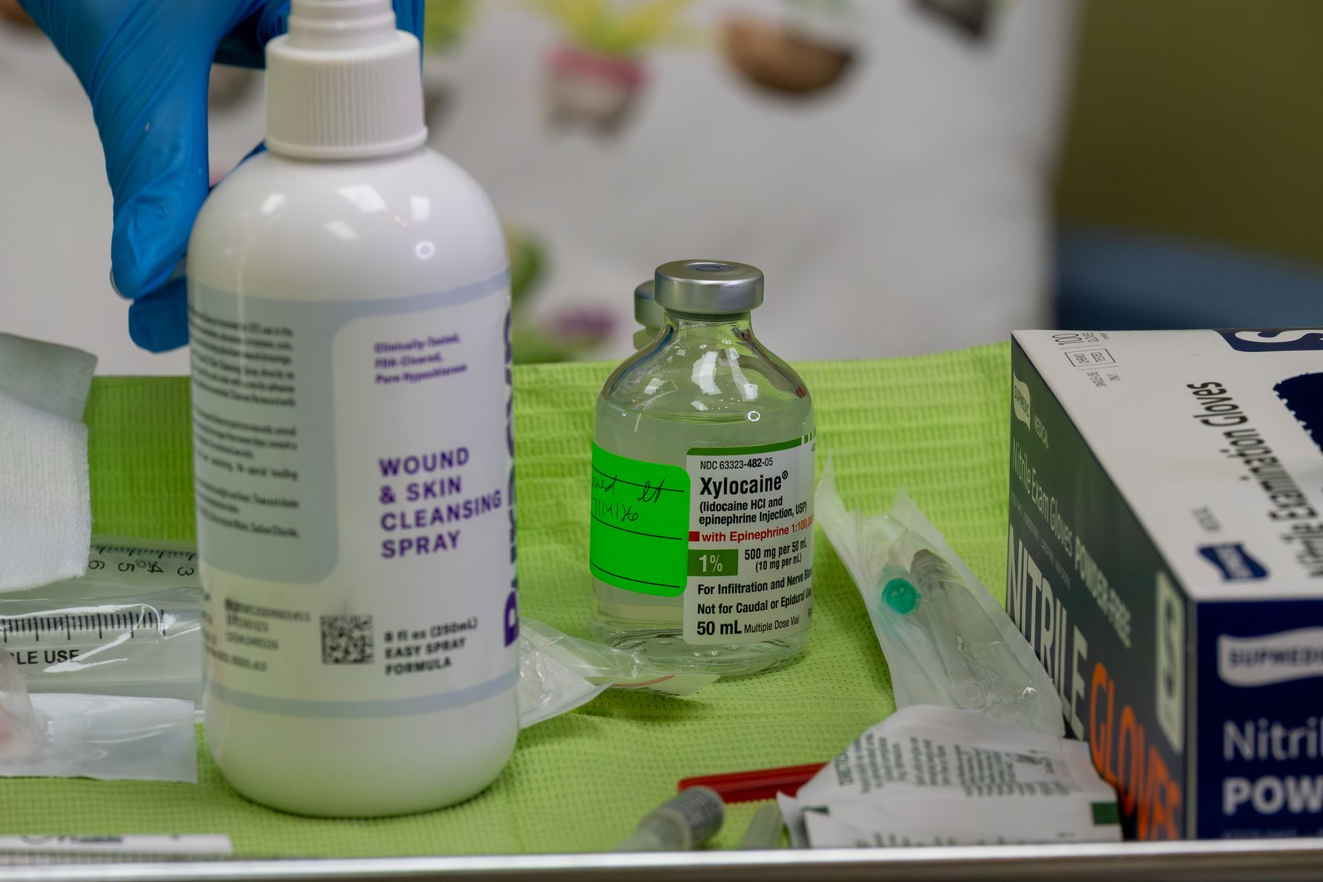 Medical supplies on a table, including a wound cleansing spray bottle and a small vial, beside gloves and gauze.